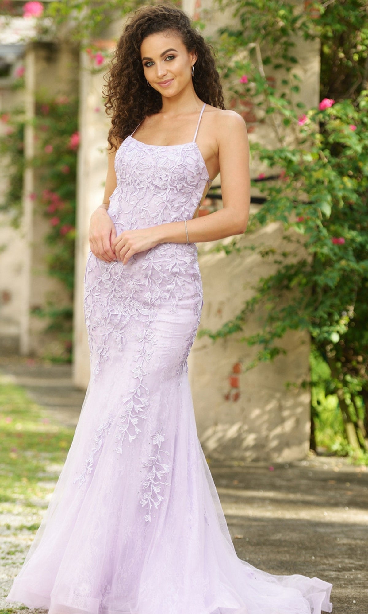 A woman stands outdoors in the Beaded Lace Long Prom Dress Dutchess, a fitted lilac gown with thin straps and floral detailing, surrounded by greenery and flowers.